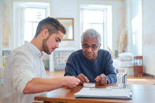 a male caregiver with a beard sitting at a wood table with an elderly man in a blue sweatshirt performing an activity at a wooden table as part of home care services.